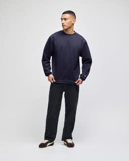 A navy blue crewneck sweatshirt on a model, paired with dark trousers and styled with casual footwear.