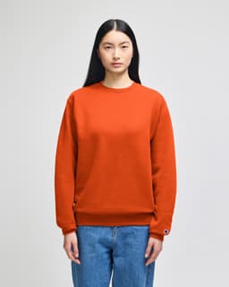 An orange crewneck sweatshirt displayed on a model, featuring a logo on one sleeve.