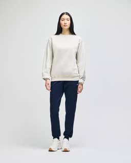 A woman stands in a light grey Champion Powerblend® adult crewneck sweatshirt and navy jogger pants, wearing white sneakers.
