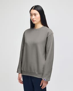 A grey Champion Powerblend® adult crewneck sweatshirt on a model, featuring a logo on the sleeve.