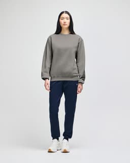 A grey crewneck sweatshirt with long sleeves, paired with dark trousers and casual shoes. The model stands against a plain background.