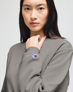 A grey Champion Powerblend® adult crewneck sweatshirt with a logo detail on the sleeve, displayed by a model.
