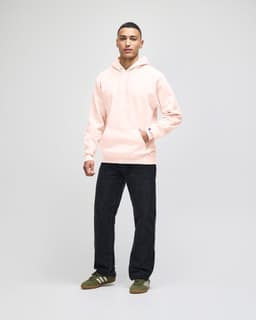 A man stands wearing a beige hooded sweatshirt and dark jeans, with sneakers. The sweatshirt has a logo.