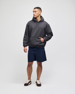 A model wears a grey hooded sweatshirt paired with navy shorts and shoes. The sweatshirt has a front pocket and a logo.