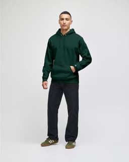 A man stands wearing an olive green pullover hooded sweatshirt and navy blue pants, with a logo visible on the hoodie.