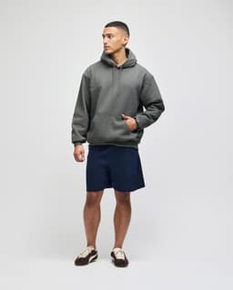 An adult pullover hooded sweatshirt in green, paired with navy shorts. The model stands confidently with one hand in the pocket.