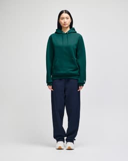 A green hooded sweatshirt with a front pocket, paired with navy trousers, displayed on a model.