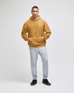 A hooded sweatshirt in a warm mustard colour paired with light grey sweatpants. The model is standing confidently.
