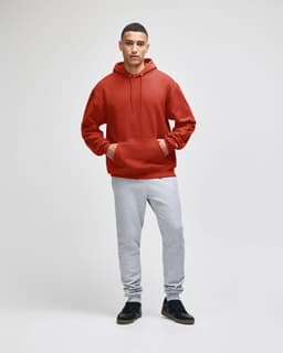 A man wearing a red pullover hooded sweatshirt and grey sweatpants, standing against a plain background.