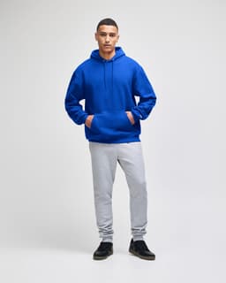 A blue pullover hooded sweatshirt with a front pocket, paired with grey sweatpants. The model stands confidently.