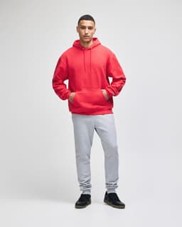 A red pullover hooded sweatshirt with a front pocket, worn by a model with navy shorts. Features a logo.