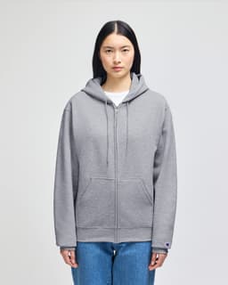 A grey full-zip hooded sweatshirt with a hood and a logo on the sleeve, styled on a model.