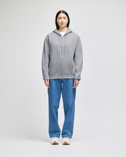 A grey full-zip hooded sweatshirt displayed on a model, paired with blue jeans. The sweatshirt features a logo.