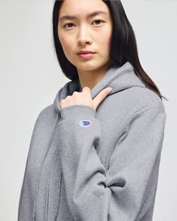 A woman wearing a grey Champion Powerblend® adult full-zip hooded sweatshirt, featuring a logo on the sleeve.