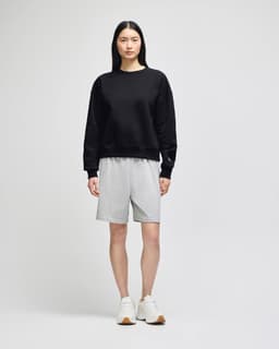 A black crewneck sweatshirt worn with grey shorts, paired with white sneakers, featuring branding.