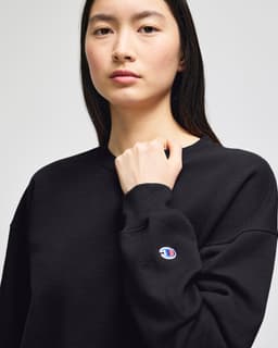 A black women's crewneck sweatshirt with a logo on the sleeve. The model is posing with a serious expression.