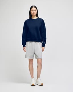 A navy crewneck sweatshirt worn with grey shorts and white sneakers, featuring a logo on the sweatshirt.