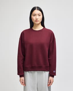 A maroon women's crewneck sweatshirt with a relaxed fit and a logo on the sleeve, paired with grey sweatpants.