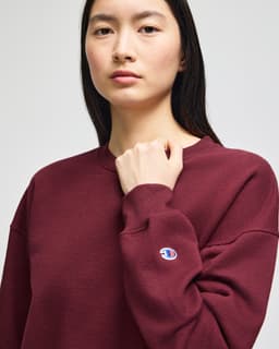 A burgundy women's crewneck sweatshirt featuring a logo on the sleeve. The model is standing and looking forward.
