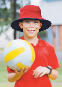 A red and navy reversible bucket hat worn by a smiling boy holding a volleyball, set outdoors.