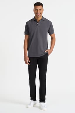 A man stands wearing a dark grey men's polo shirt, complemented by black pants and white shoes. The shirt features a logo.