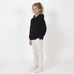 A young girl stands wearing a black hooded sweatshirt and white pants, with a light background. The hoodie has a logo.