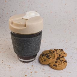 A reusable coffee cup in grey with a lid, featuring a felt band, accompanied by chocolate chip cookies.