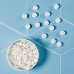 A white bowl filled with round white mints, with several mints scattered around on a blue surface.