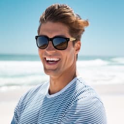 Bamboo sunglasses with dark lenses, worn by a smiling man in a striped shirt, set against a beach backdrop.