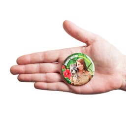 A hand holding a 57mm round badge featuring a vibrant design and a logo. The badge includes a colourful background.