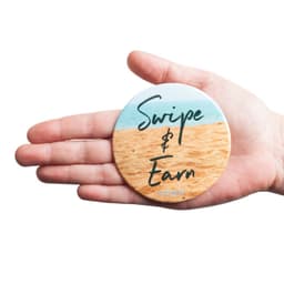 A round badge held in a hand, featuring a sandy beach design with a light blue background and green text.