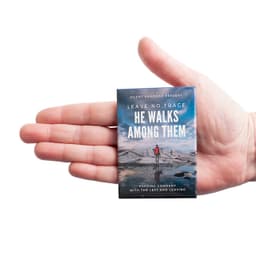 A hand holds a rectangular badge featuring a scenic landscape with a person standing by water, along with a logo.