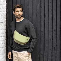 A waist bag in light green worn across a man's chest, with a casual grey sweater and beige pants, against a dark wooden background.