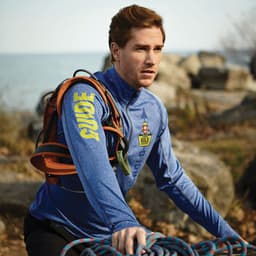 A man wearing a blue quarter zip jumper, with a logo and harness, is outdoors near water and rocks.