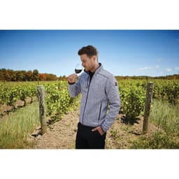 A man in a grey knit jacket poses with a wine glass in a vineyard setting, with green vines and blue sky in the background.
