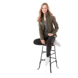 A woman wearing a green Peyto softshell jacket, seated on a stool, styled with black pants and white shoes.