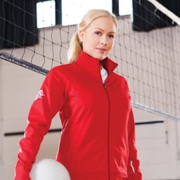 A woman in a red knit jacket stands against a volleyball net, holding a ball. The jacket features a logo.