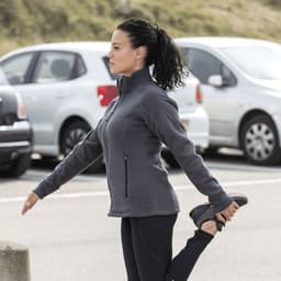 A woman stretches while wearing a grey polar fleece jacket, with parked cars in the background. The jacket features a logo.