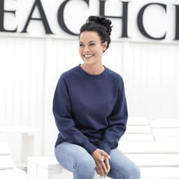 A woman sits wearing a navy blue fleece crew jumper, paired with light blue jeans, smiling against a neutral background.