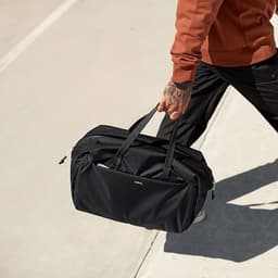 A black duffle bag with sleek lines, being held by a person wearing a brown sweater and black pants. The bag has a logo.