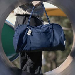 A navy duffle bag with handles is held by a person, viewed through a circular frame. The bag has a tag and a logo.