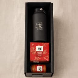 A black drink bottle, a red box of Christmas pudding, and a jar of sauce, all in a black hamper.