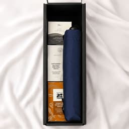 A navy umbrella is nestled in a box with treats like cinnamon balls and other snacks. The box has a sleek design and logo.