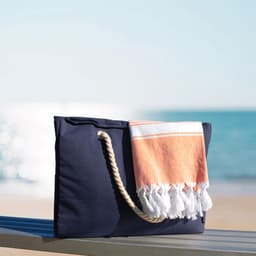 A navy waterproof beach bag with a woven handle and a folded orange and white towel draped over it.