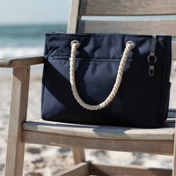 A navy waterproof beach bag with rope handles rests on a wooden chair by the shore. Features a logo.