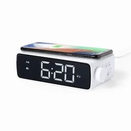 A digital alarm clock in white with a large display showing the time and a smartphone resting on top for charging.