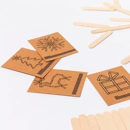 A collection of brown craft cards featuring black line drawings of a snowflake, tree, cactus, and gift, alongside wooden sticks.