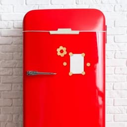 A red retro fridge with a shiny surface, featuring a magnetic note board and decorative magnets on a white brick wall.