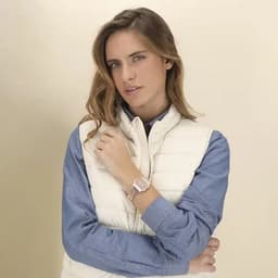 A watch with a silver band is worn by a woman in a light vest over a blue shirt, posing against a neutral background.