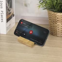 A mobile phone displaying a video is propped up on a wooden stand beside a potted plant and a book.